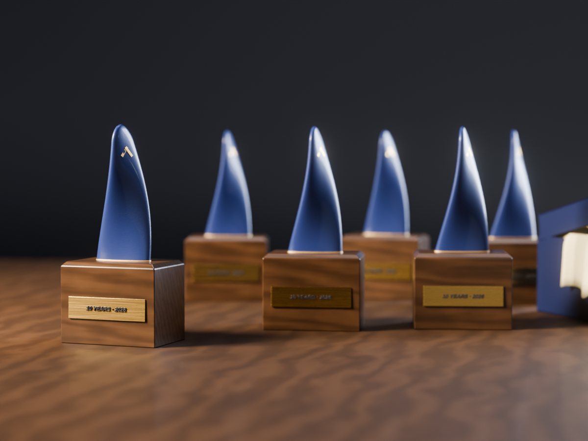 Concept render of six identical navy desk awards on walnut plinths with brass plates reading 10 YEARS 2026, one in focus at the front, on a walnut table in a dark studio