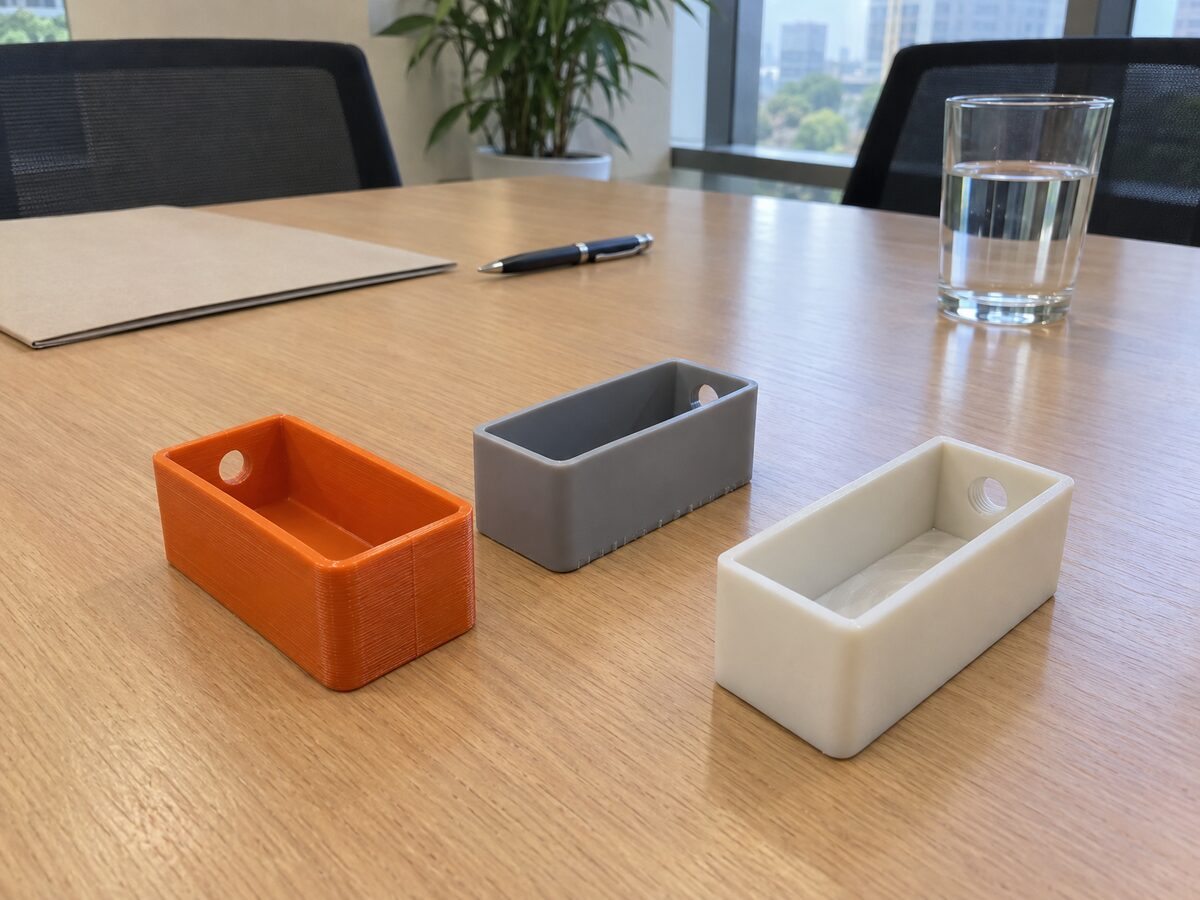 Three palm-sized open housings on an office conference table: orange FDM with layer lines, smooth grey SLA resin, and off-white machined plastic with a tapped hole