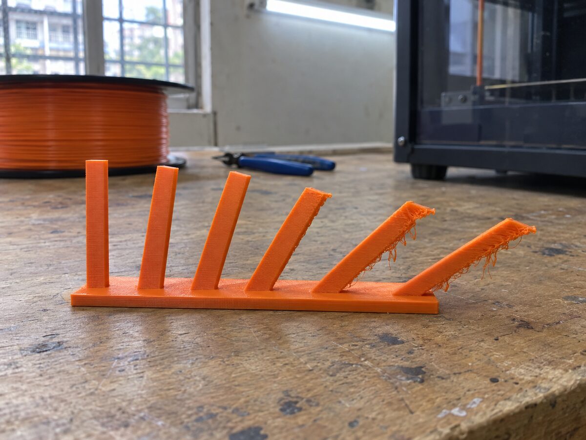 An orange overhang test print on a worn wooden workbench: fins lean out at steeper angles, clean on the left and drooping with rough undersides on the right