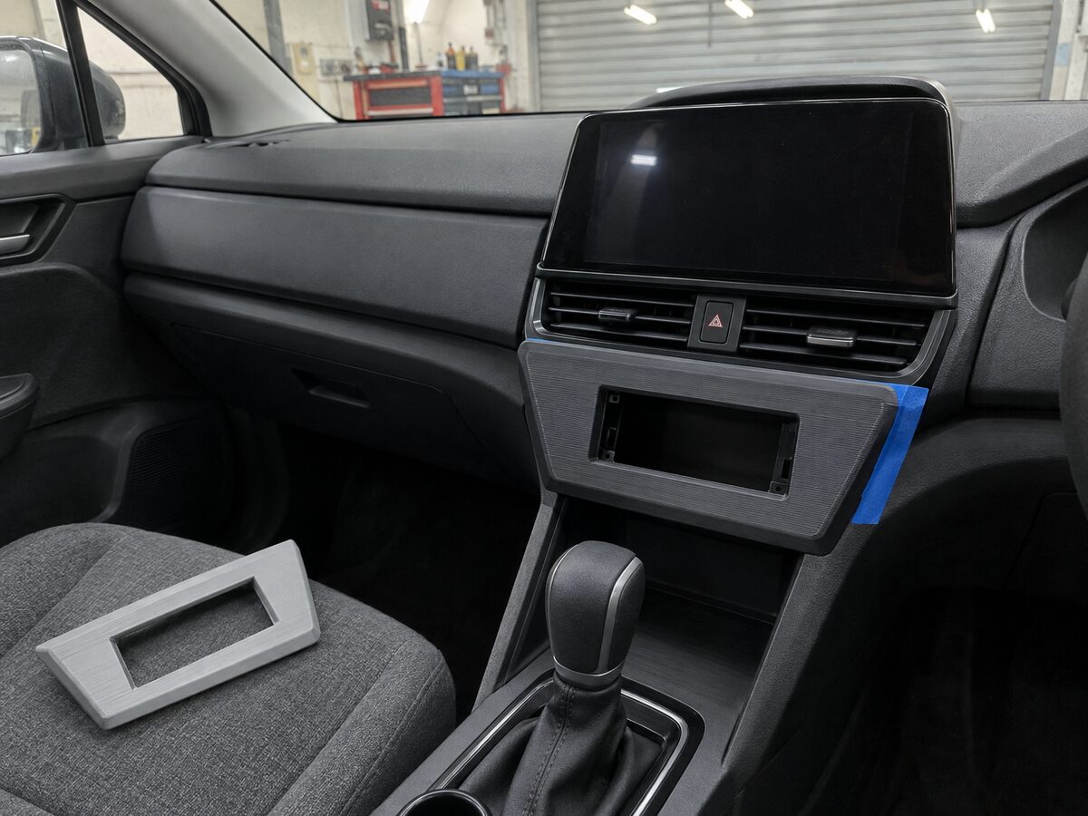A charcoal printed prototype trim bezel test-fitted in a car's centre console, with a light-grey version lying on the passenger seat