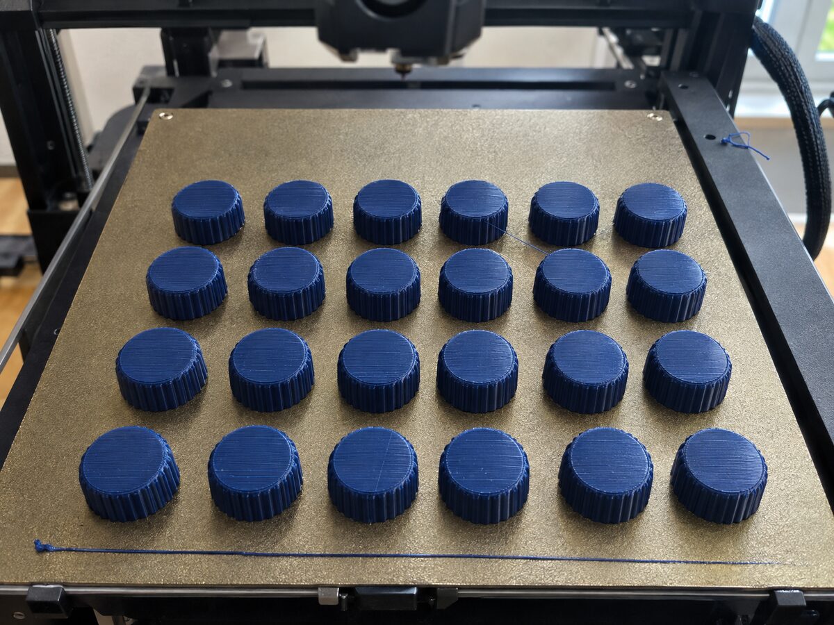 Open desktop FDM printer whose textured gold build plate is filled with rows of identical navy PETG control knobs, freshly printed