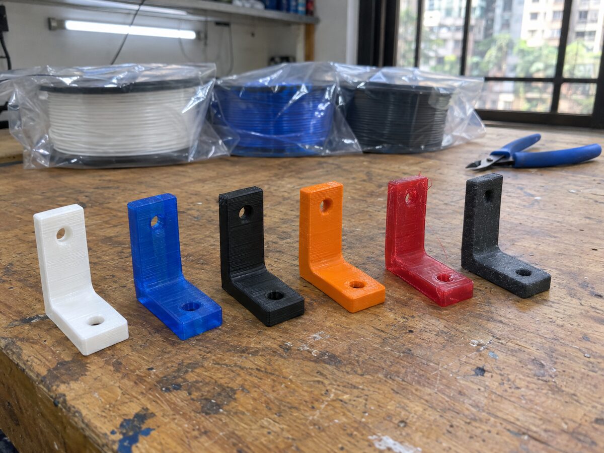 Six identical small test brackets printed in PLA, PETG, ABS, ASA, TPU and carbon-fibre nylon, standing in a row on a workshop bench