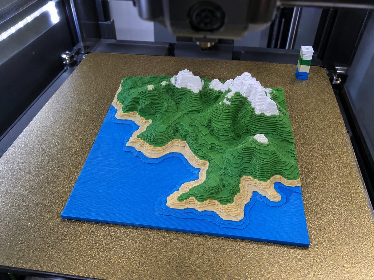 A square terrain relief tile printed in four colours, blue, beige, green and white, on a gold build plate inside an enclosed printer, with a purge tower beside it