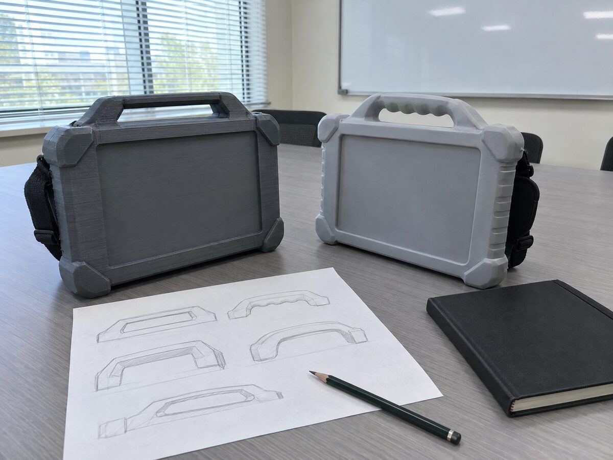 Two full-size printed mock-ups of a rugged field tablet with bumpers and carry handles, one raw grey print and one primed, on a meeting-room table