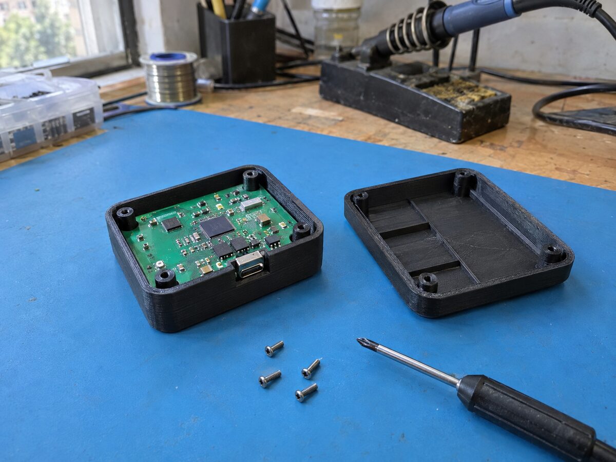 Palm-sized matte black 3D-printed enclosure base with a circuit board seated on its screw bosses and the lid beside it, on an electronics bench