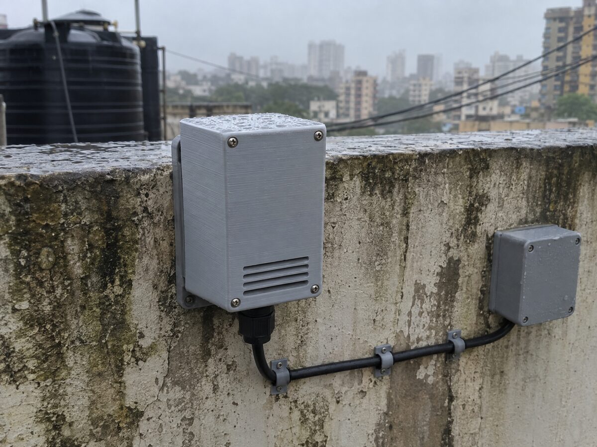 A light-grey printed sensor enclosure with vent slots and a cable gland, screwed to a damp concrete terrace wall in Mumbai during the monsoon