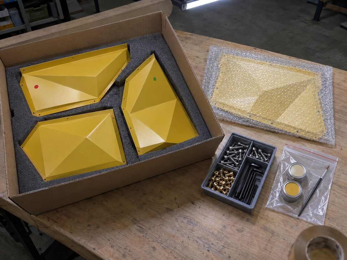Painted mustard-yellow model sections in a foam-lined carton with colour-dot tags, beside a tray of bolts and hex keys and a bag of touch-up paint tins