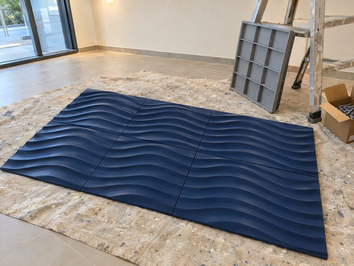 Six 3D-printed wave-relief wall tiles painted satin navy, laid out on a dust sheet in an office reception, with a seventh leaning on a ladder to show its raw grey printed back