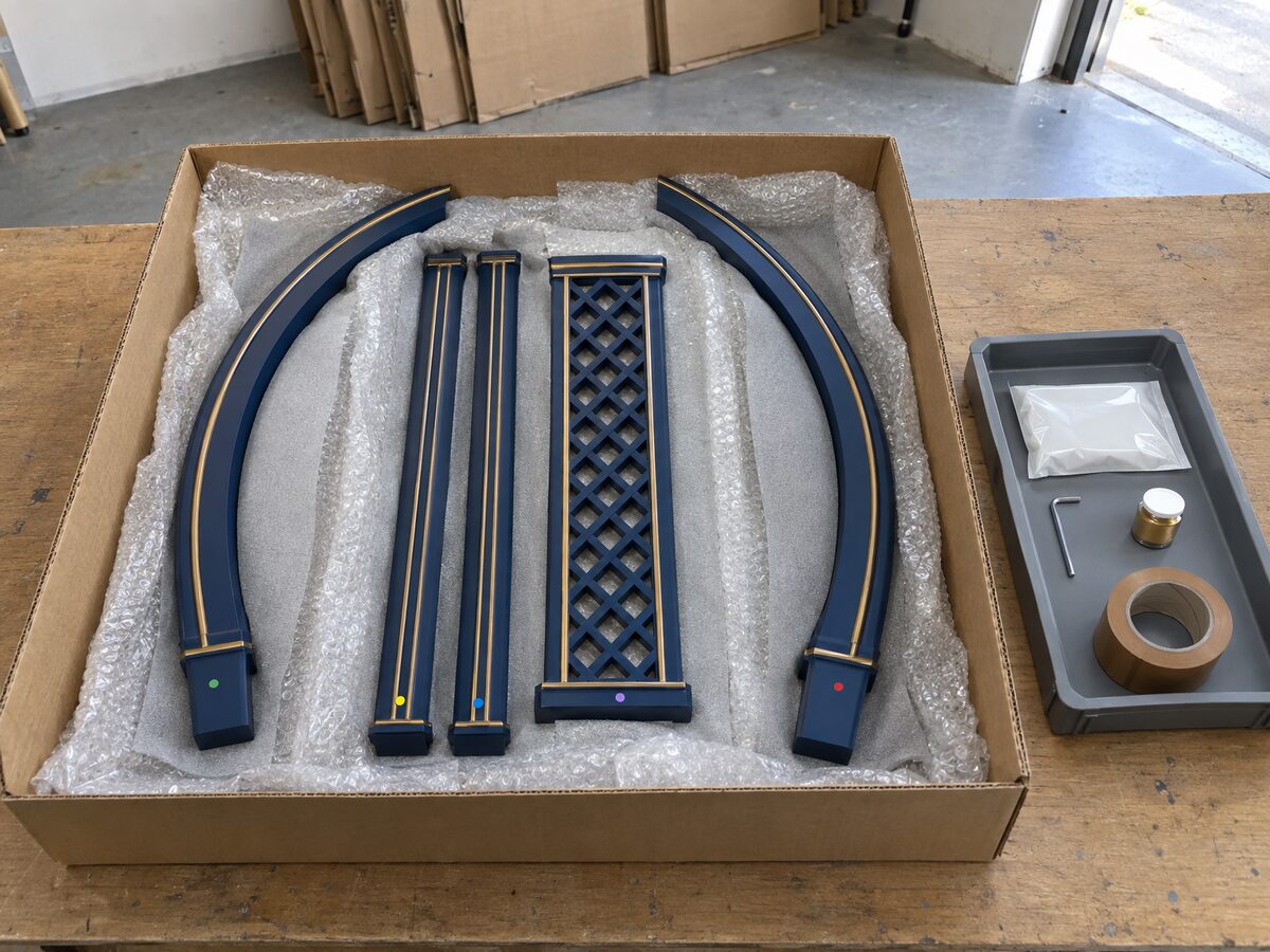 Open cardboard carton holding flat-packed navy and gold makhar arch, pillar and lattice sections in bubble wrap, beside a tray with a fixings pouch, hex key, touch-up paint and tape