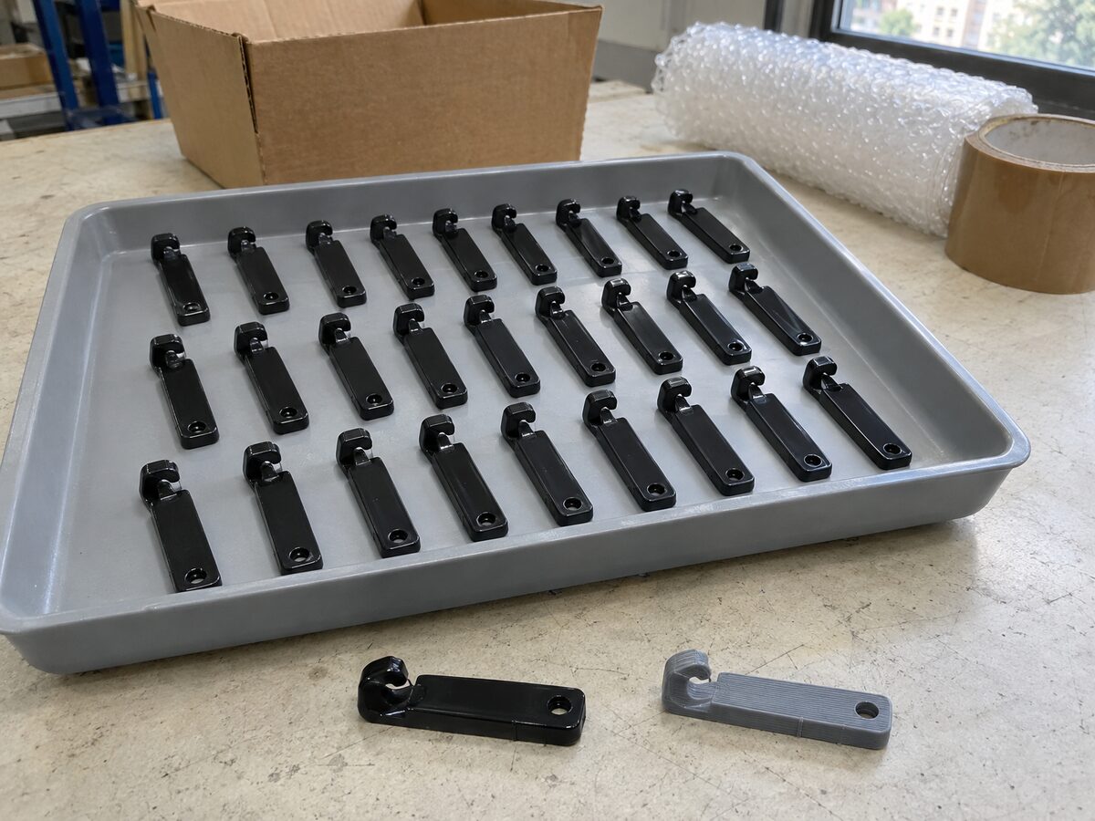 Tray of identical glossy black injection-moulded clips in rows on a packing table, one clip beside its grey 3D-printed prototype