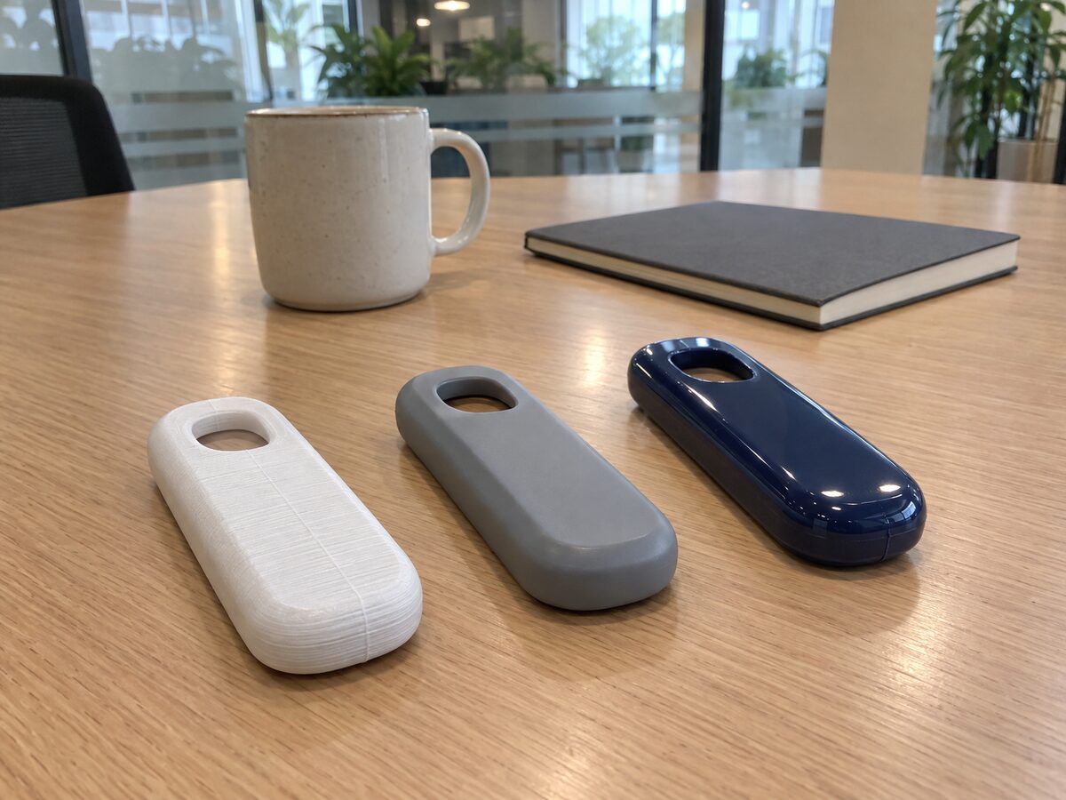 The same handheld housing three ways on an office meeting table: a white FDM print with layer lines, a smooth grey SLA print and a glossy navy moulded part